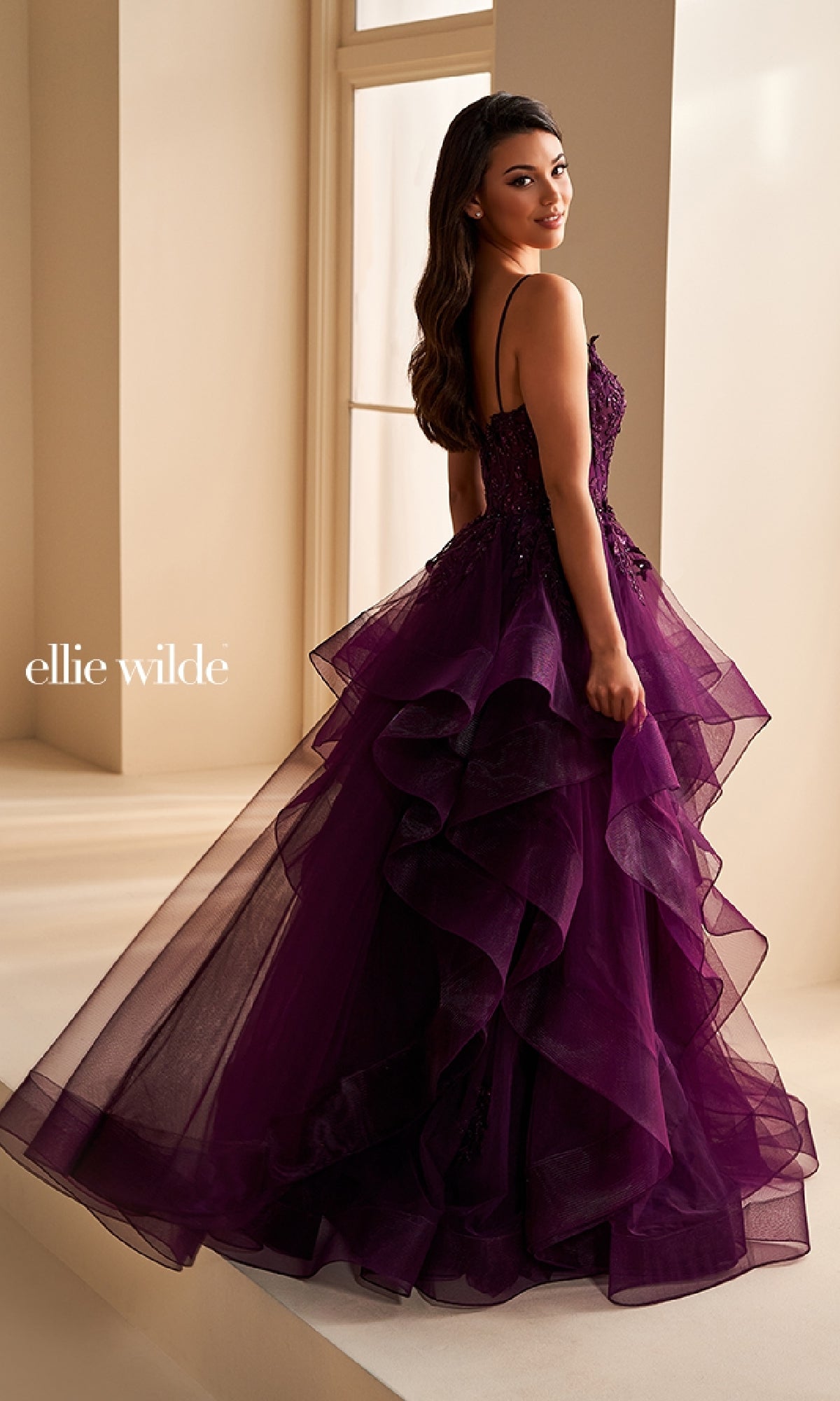 A woman in a purple, tiered tulle ball gown with spaghetti straps and floral accents stands by a window, glancing over her shoulder. She wears the stunning Ellie Wilde EW36225 long prom dress.